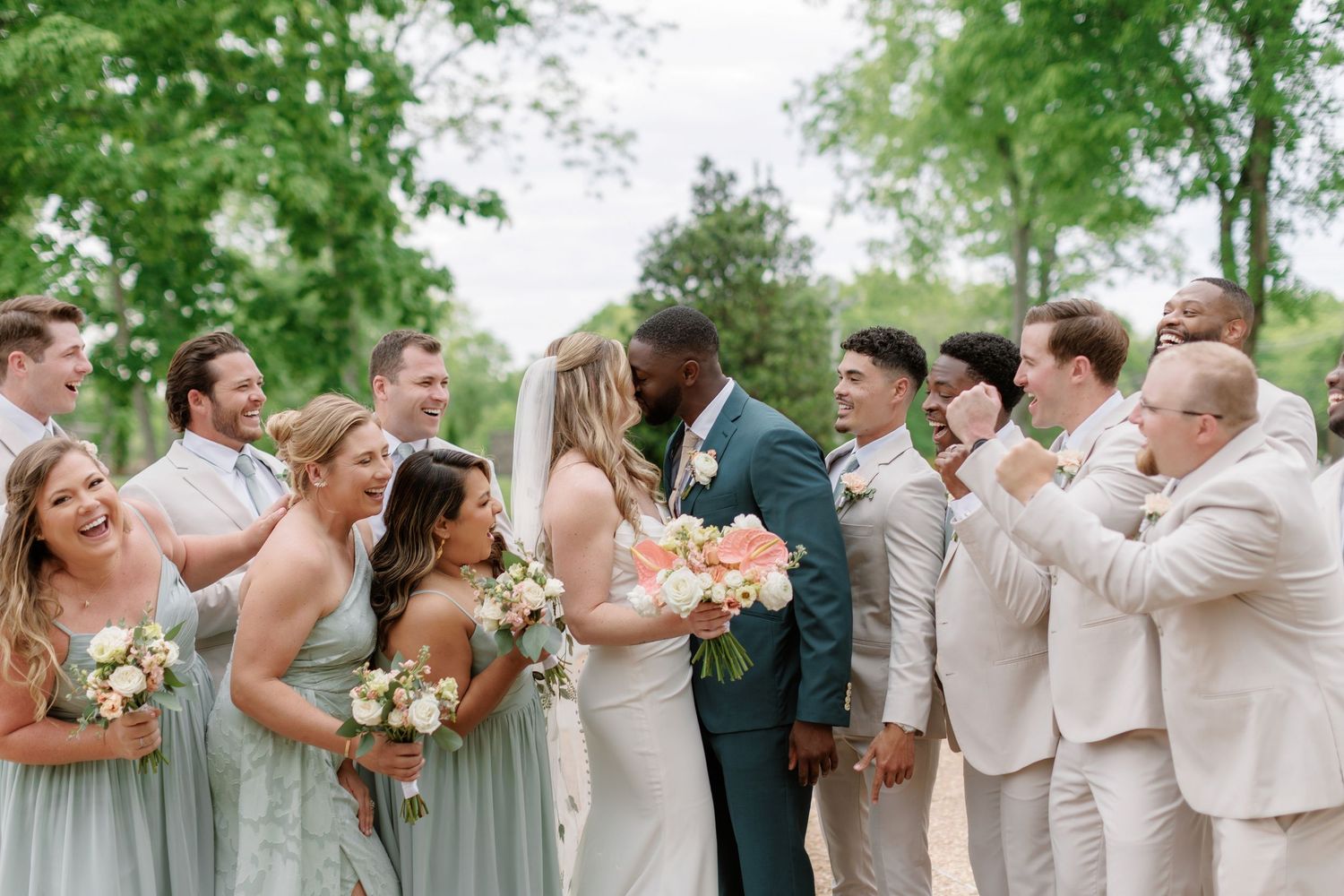 Wedding party in sage green and beige attire gathers outdoors for celebratory photo with blossoming trees in background.