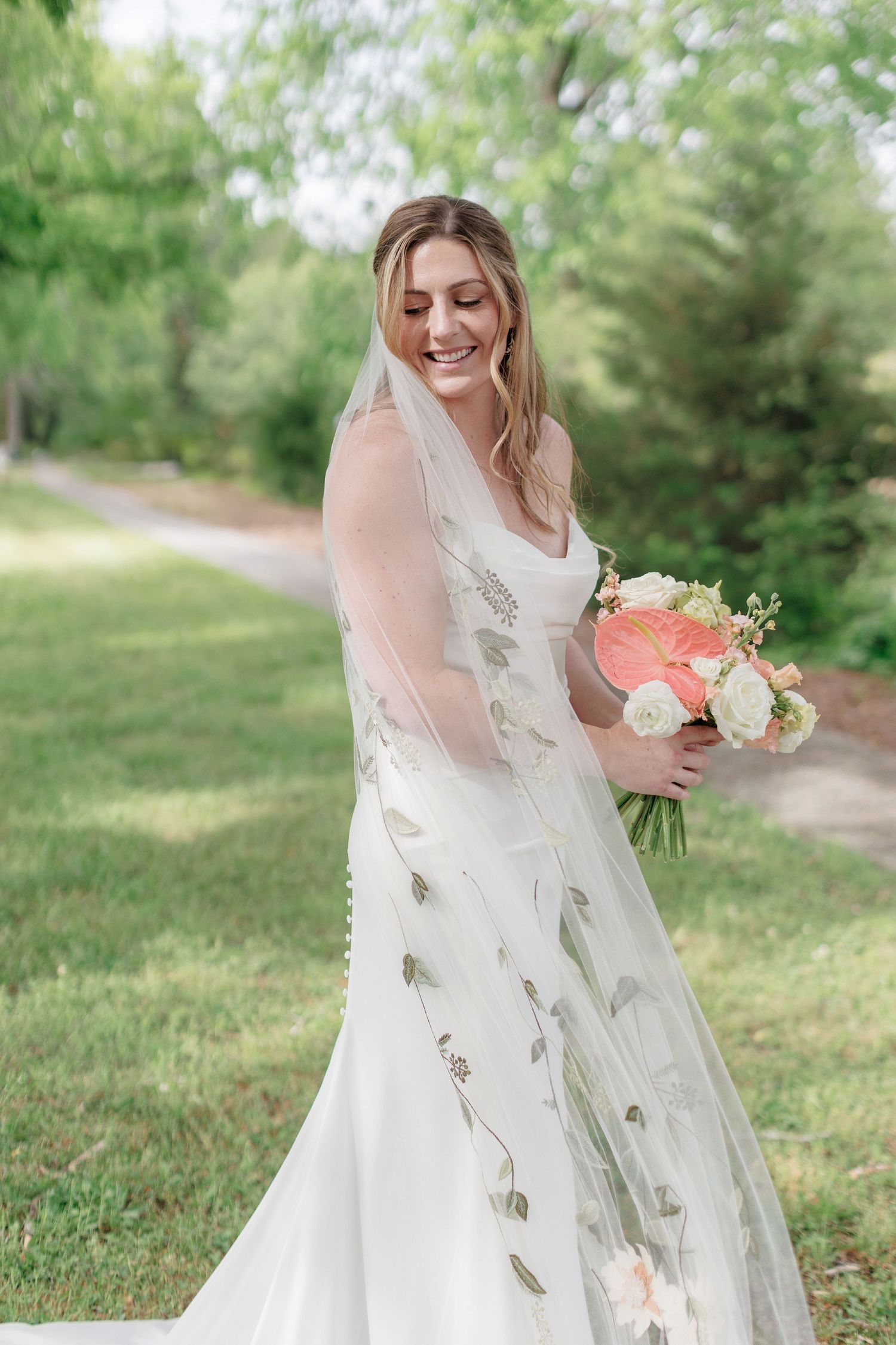 Beautiful outdoor wedding photo featuring bride in flowing white dress with delicate leaf details and coral bouquet in garden setting.