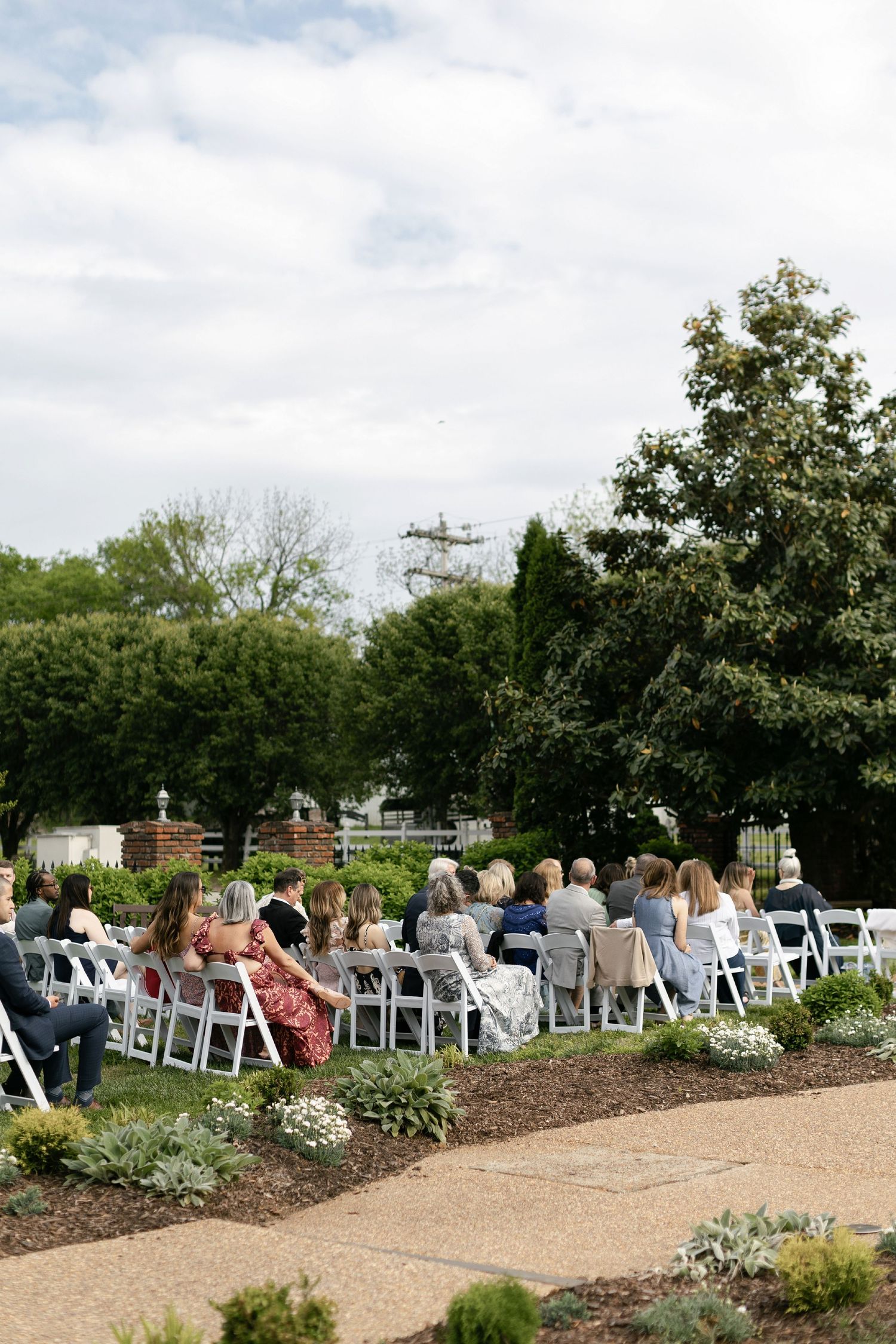 Guests seated in white folding chairs for an outdoor ceremony along a winding garden path on a cloudy day.