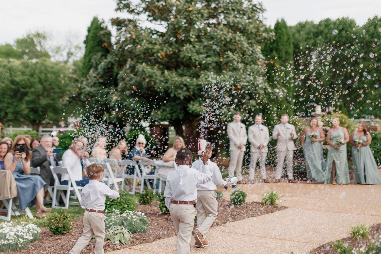 Ringbearers blow bubbles as bridal party walks along a garden path at an outdoor ceremony.