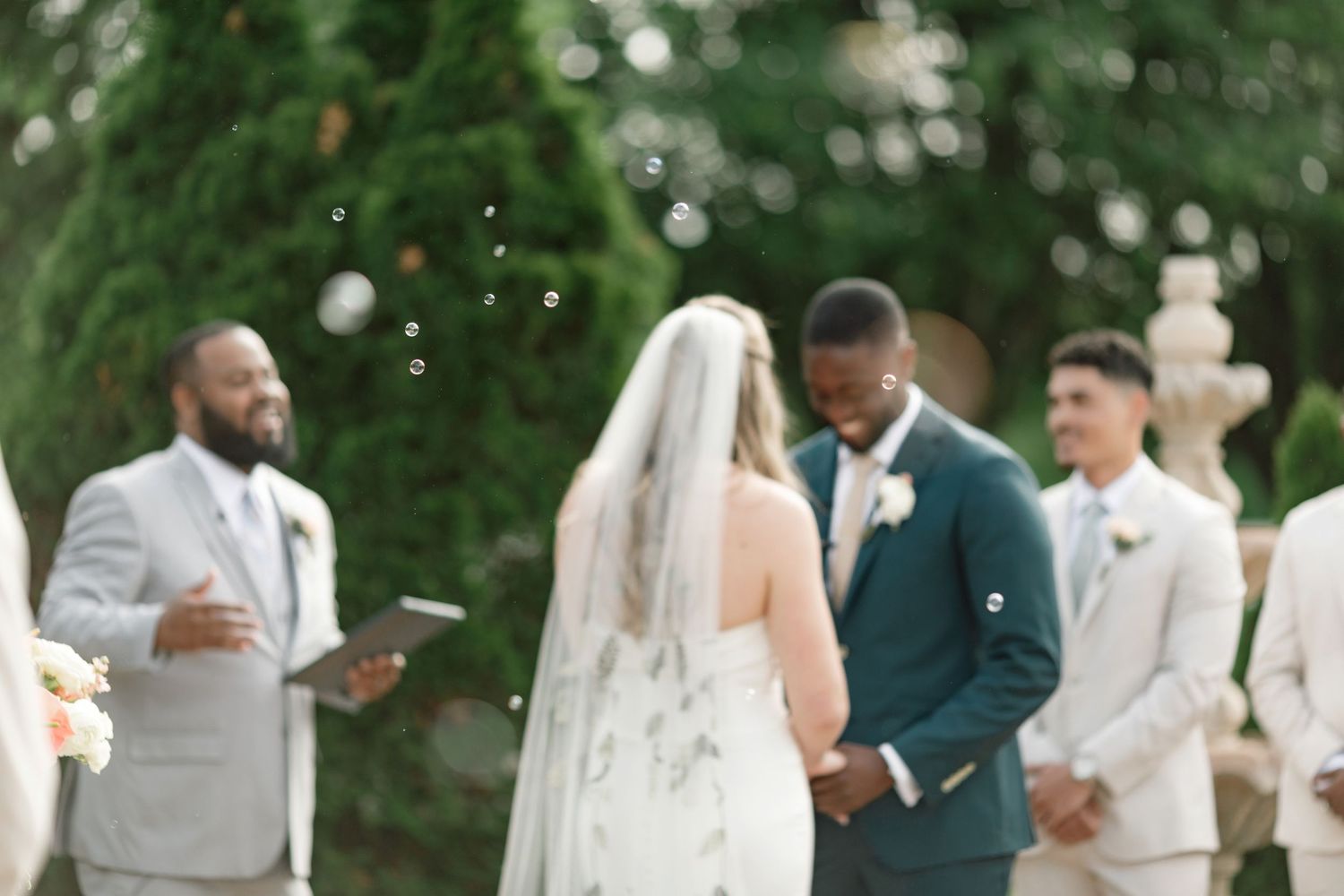 An outdoor wedding ceremony in a garden setting with wedding party members in light gray and navy suits.