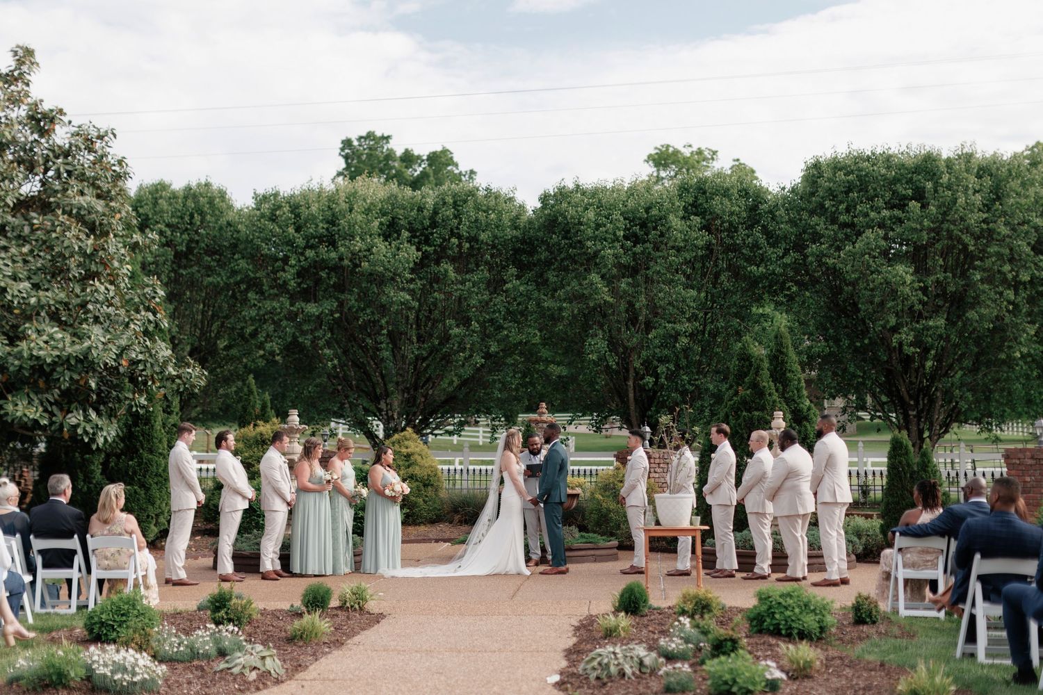 An outdoor wedding ceremony takes place in a garden setting surrounded by lush green trees at Legacy Farms near Nashville, TN.