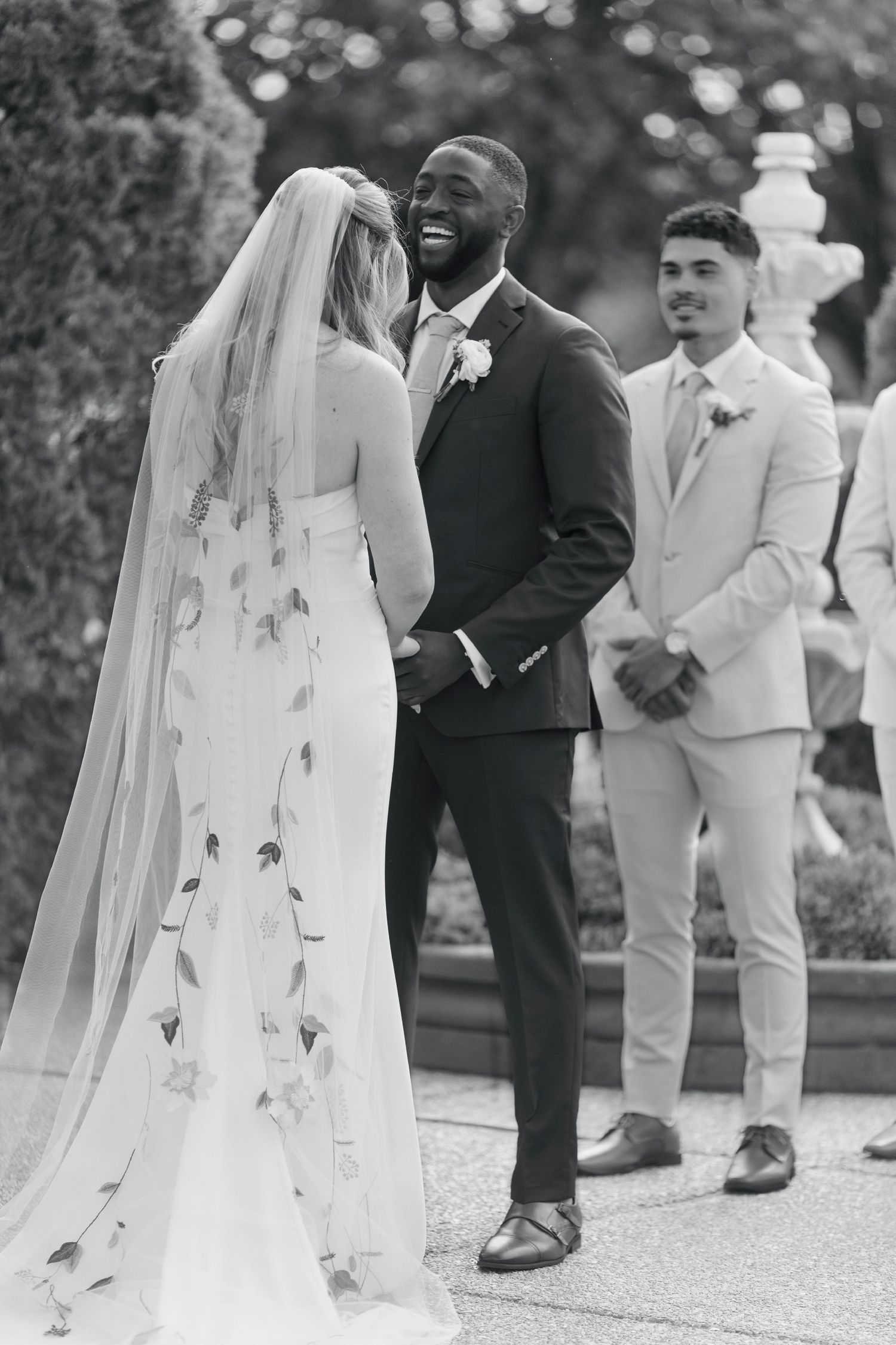 A bride in a flowing veil shares a candid laugh with someone at an outdoor garden wedding ceremony.