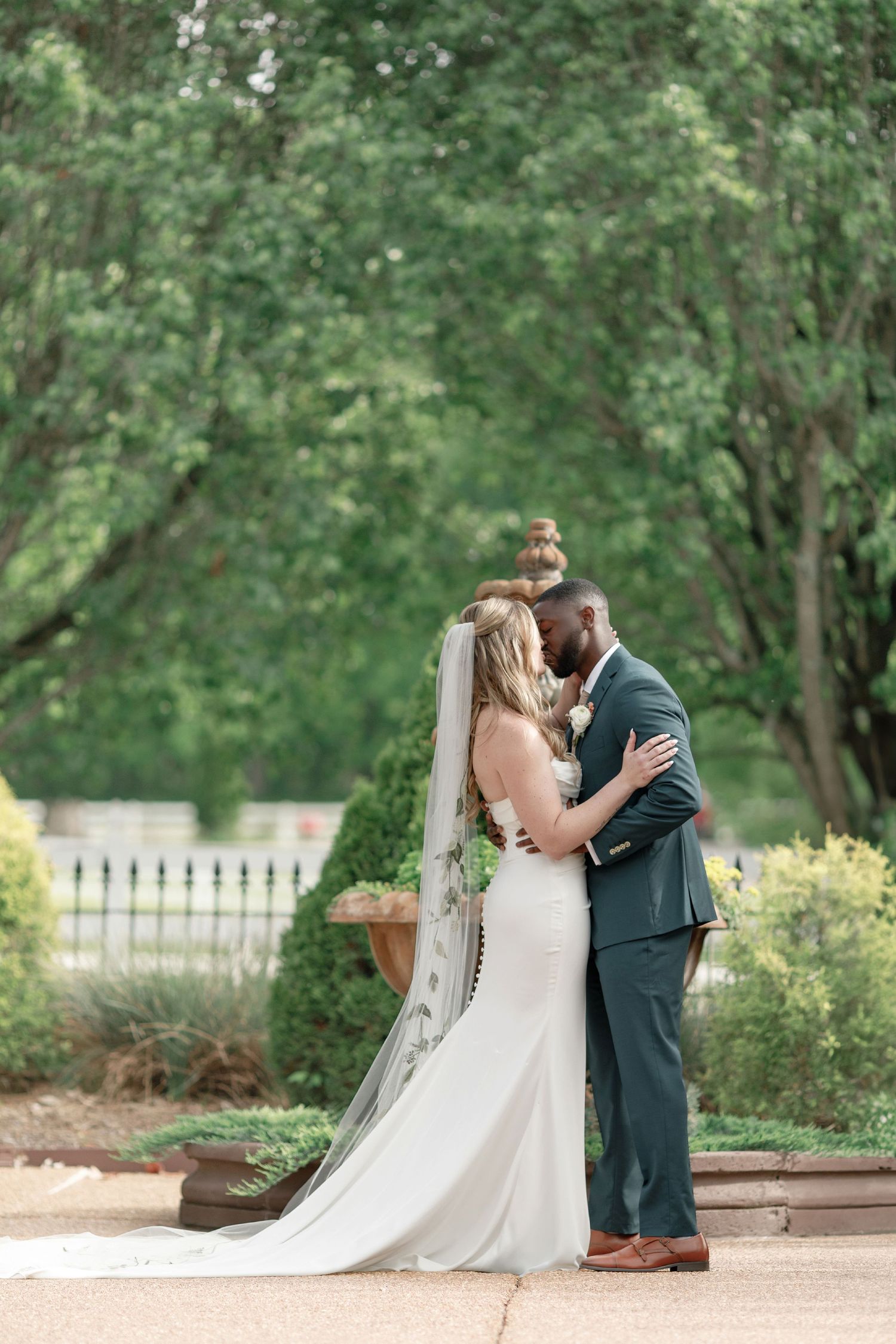 A romantic outdoor wedding portrait featuring a bride in white gown with flowing veil embracing groom in gray suit.