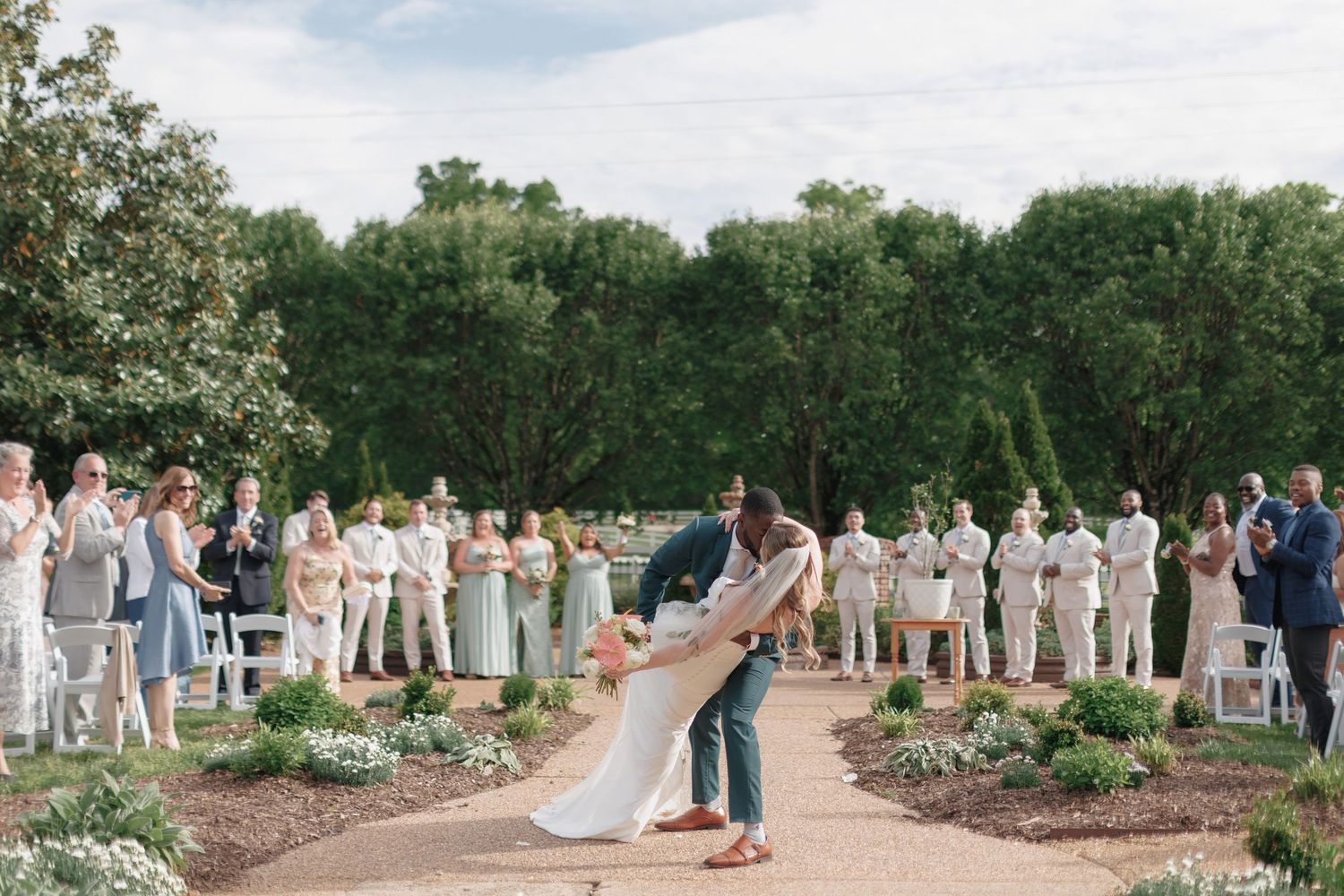 A couple shares their first dance surrounded by their wedding party in a lush garden setting.