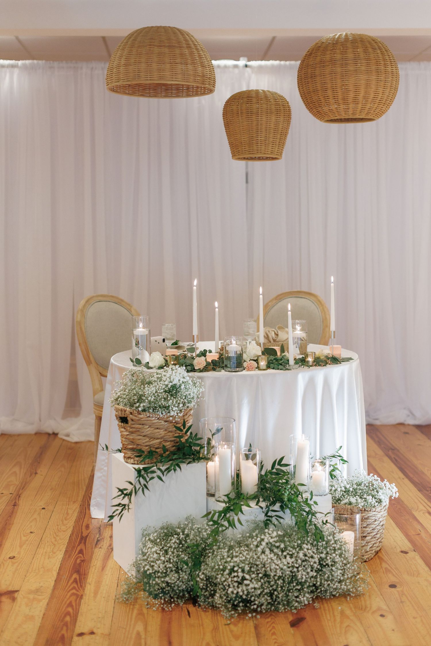 Elegant table setting with basket pendant lights, white linens, candles, and baby's breath floral arrangements on wooden floor.