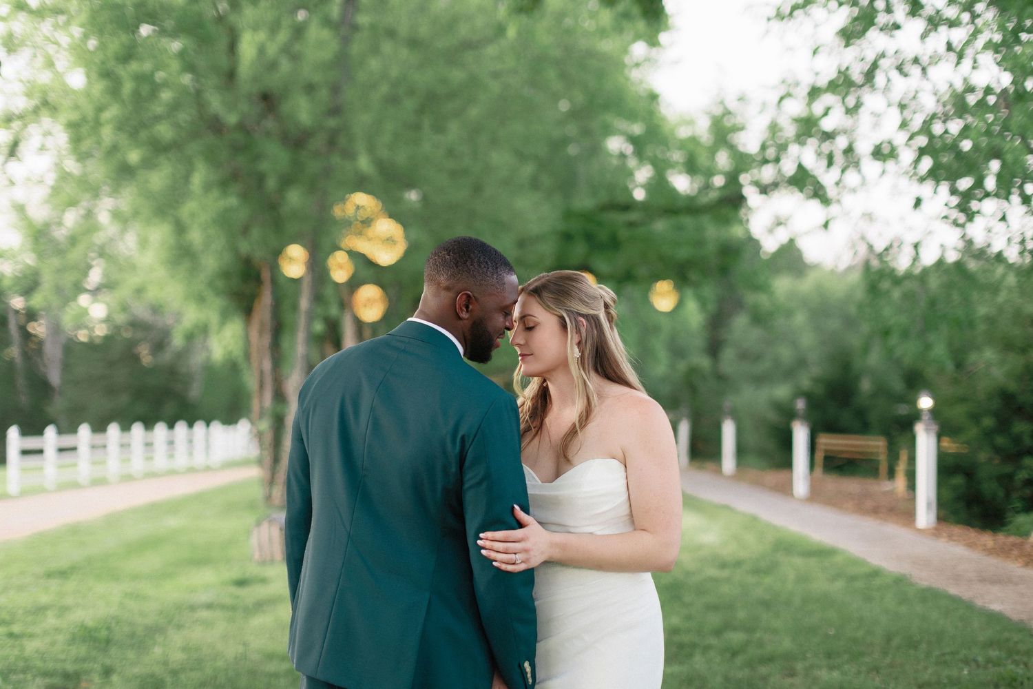 A romantic couple shares an intimate moment outdoors on a path with trees and white fence in the background.