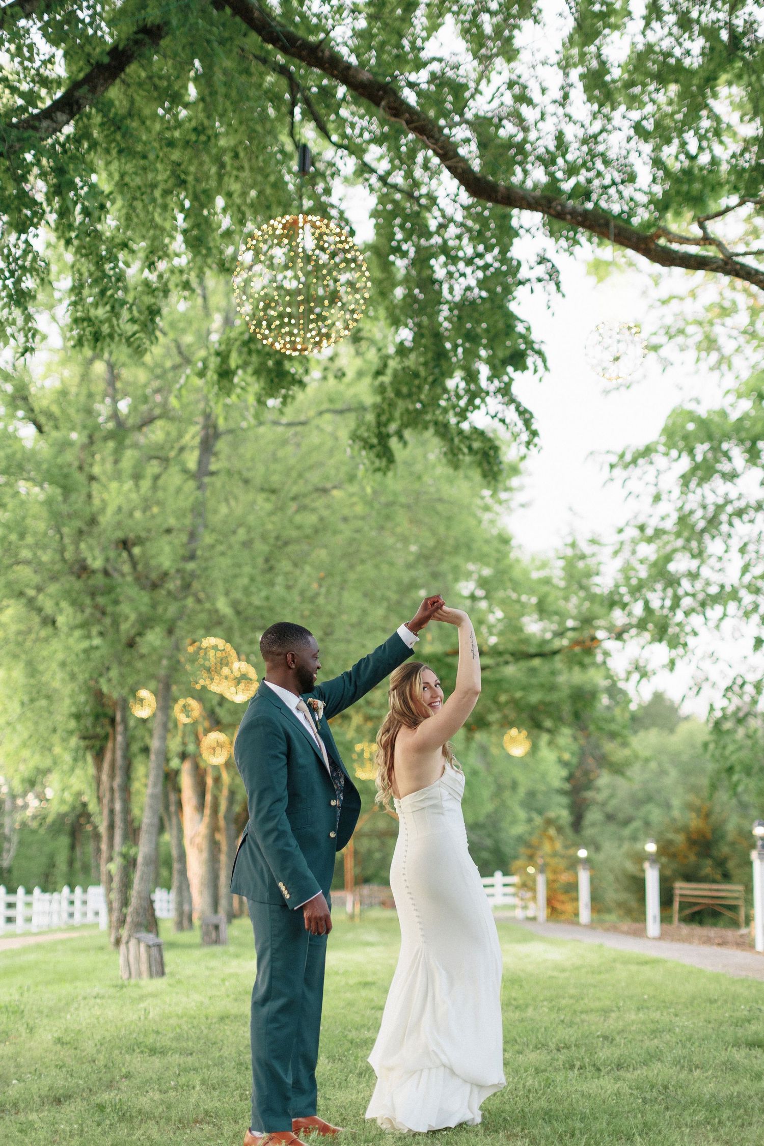 A couple dances joyfully on a lush green lawn surrounded by trees and hanging lanterns during an outdoor celebration.