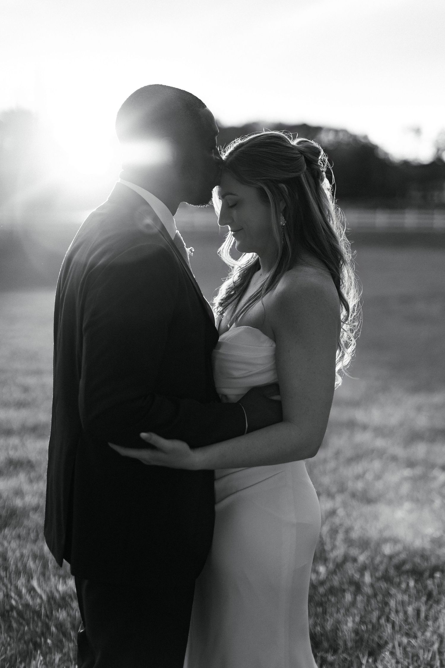 A romantic black and white wedding photo showing a couple embracing in soft sunlight on a grassy field.