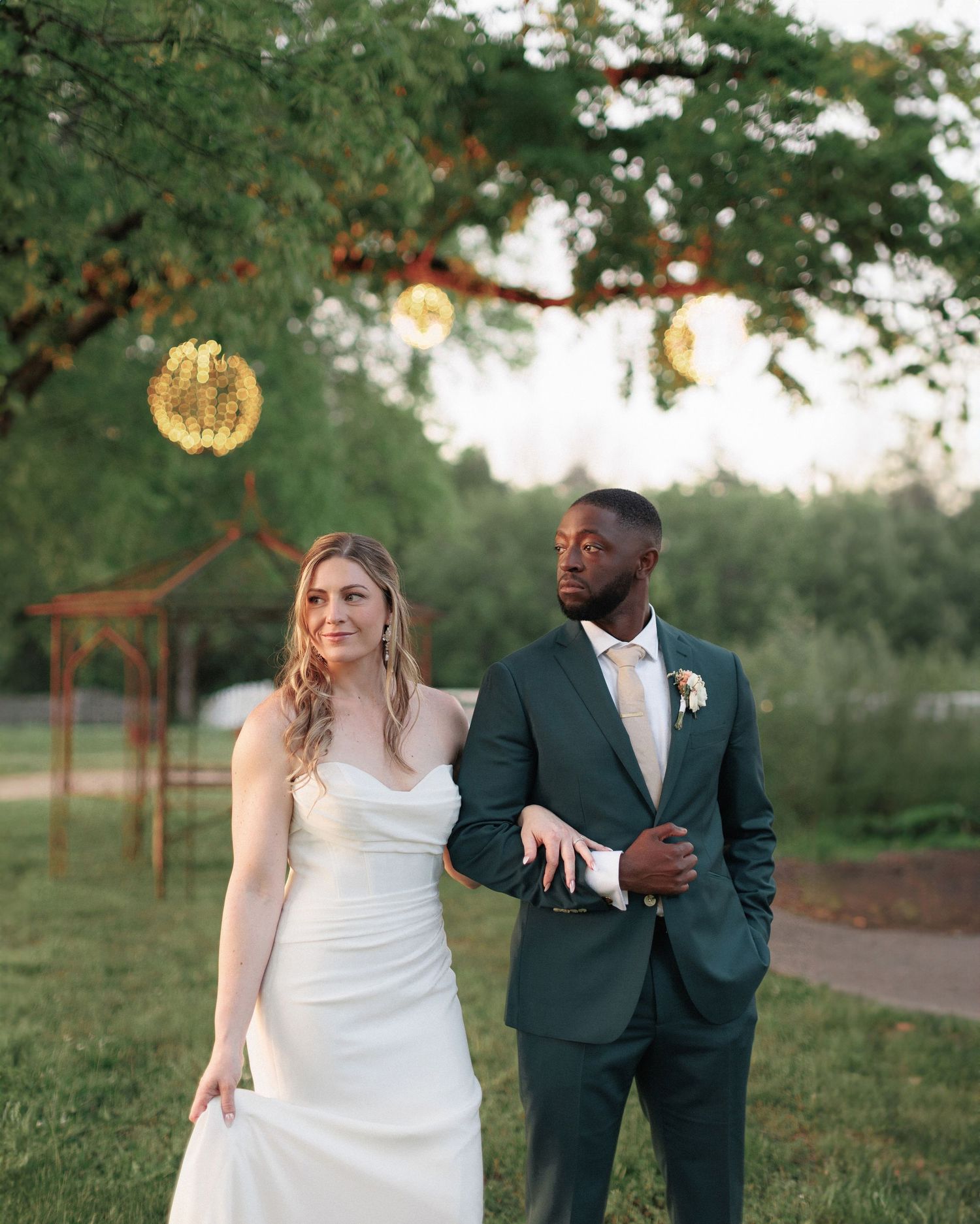 A couple in wedding attire stands outdoors at sunset with glowing decorative lights and greenery in the background at Legacy Farms.