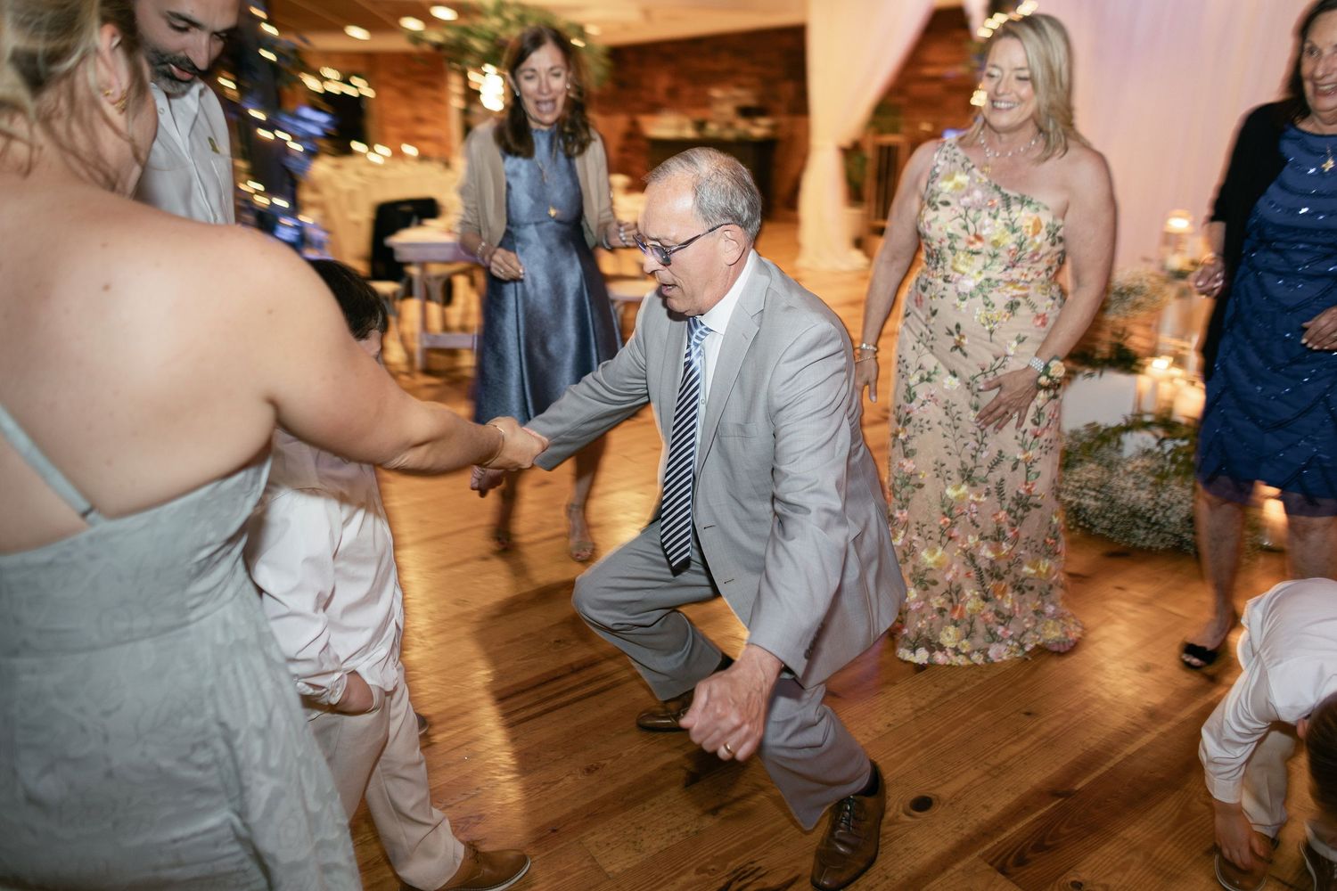Wedding guests dancing and celebrating on a wooden dance floor at a reception venue with warm lighting.