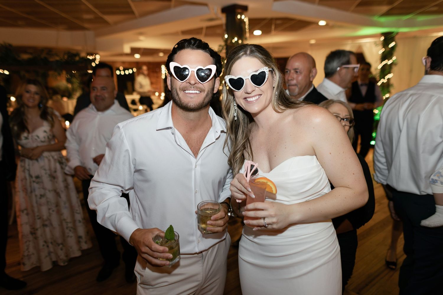Party guests celebrate on the dance floor at a reception wearing sunglasses and smiling.