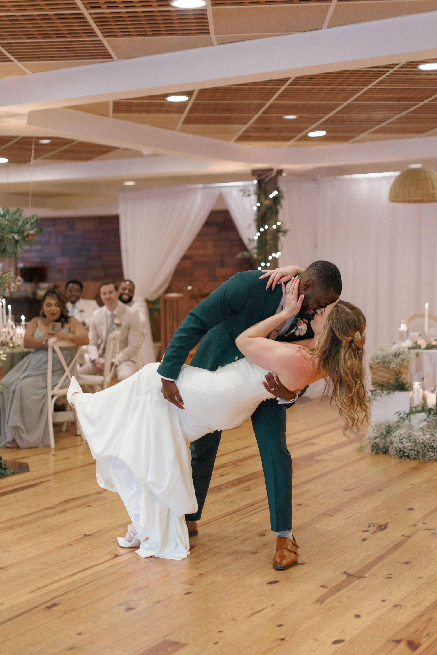 A couple shares a romantic dip during their first dance on a wooden floor at their wedding reception.