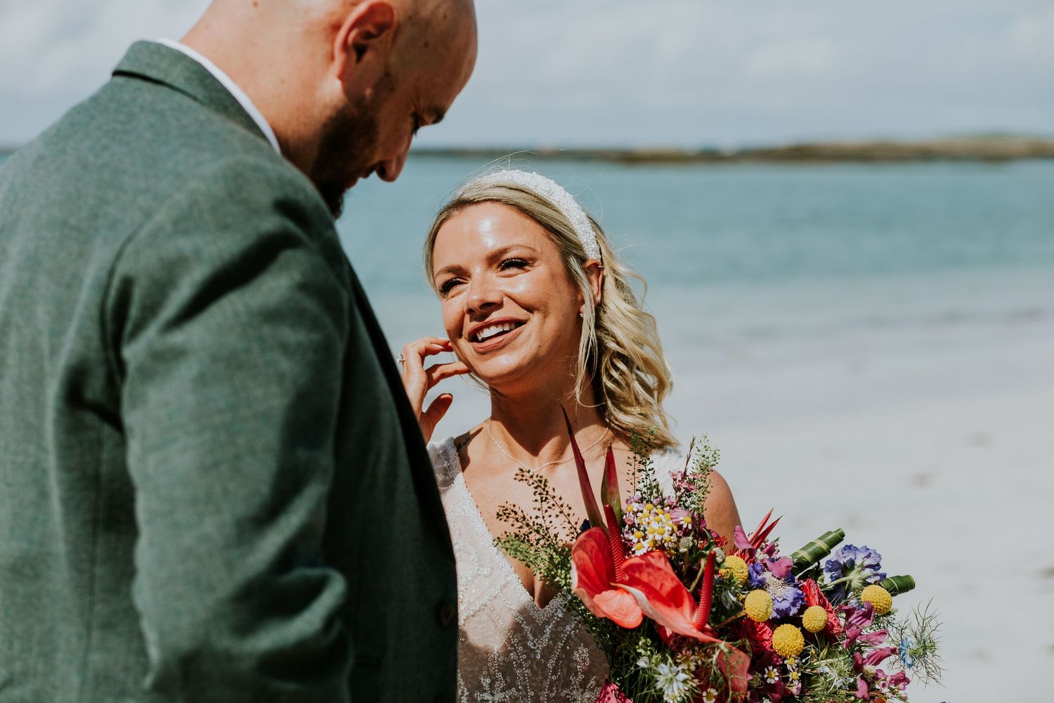 Beach wedding moment captured with vibrant floral bouquet against turquoise ocean backdrop.