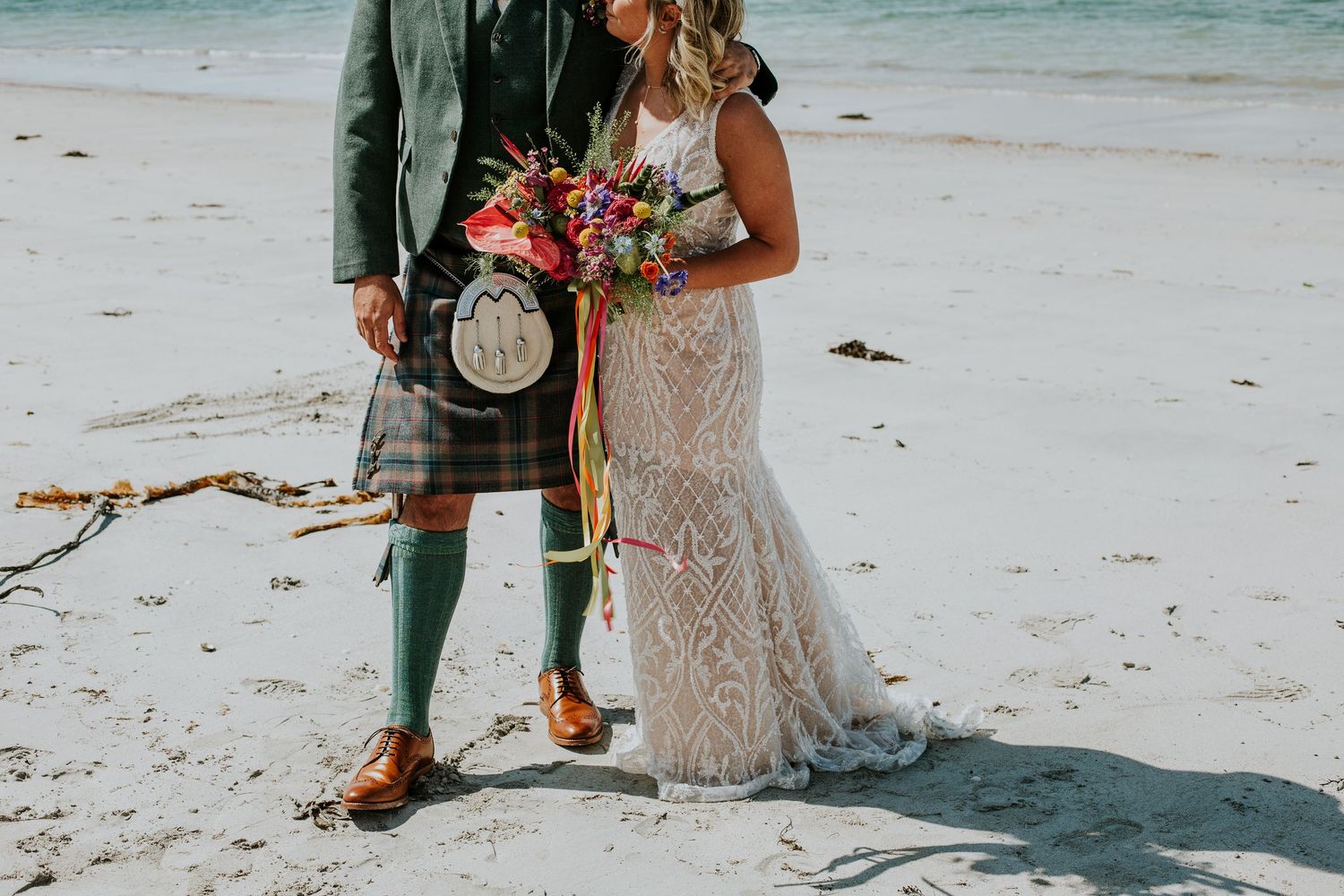 A couple in wedding attire featuring a traditional kilt and lace dress stand together on a beach.