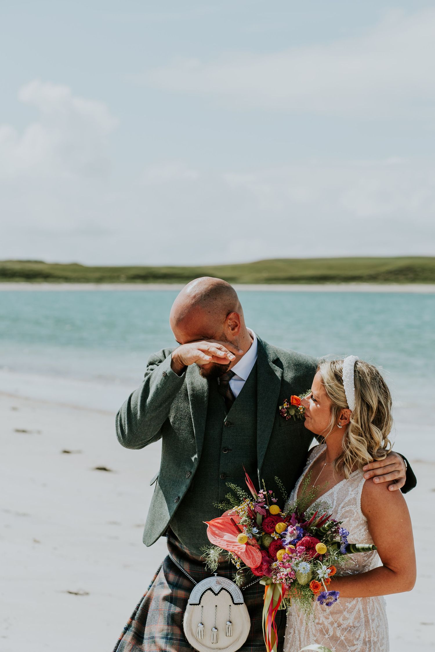 A wedding couple shares an emotional moment on a scenic beach with turquoise waters and cloudy skies.