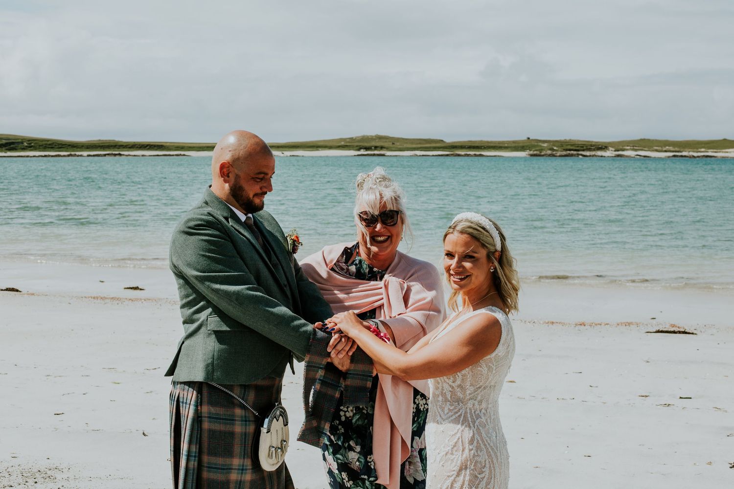 Three people stand together in a ceremony on a scenic beach with turquoise water in the background.