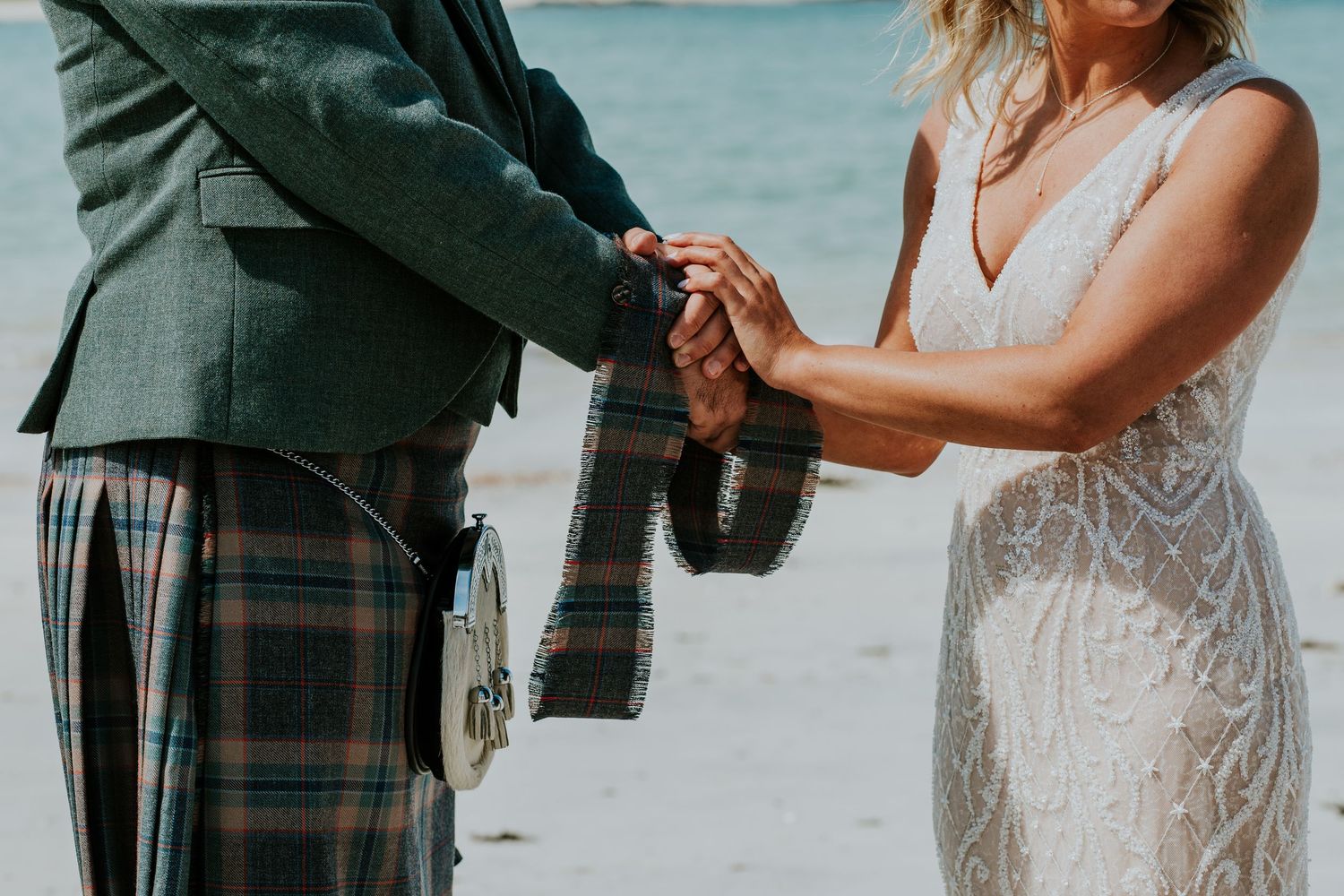 A couple holds hands during their beach wedding ceremony, with the groom wearing a traditional Scottish kilt.