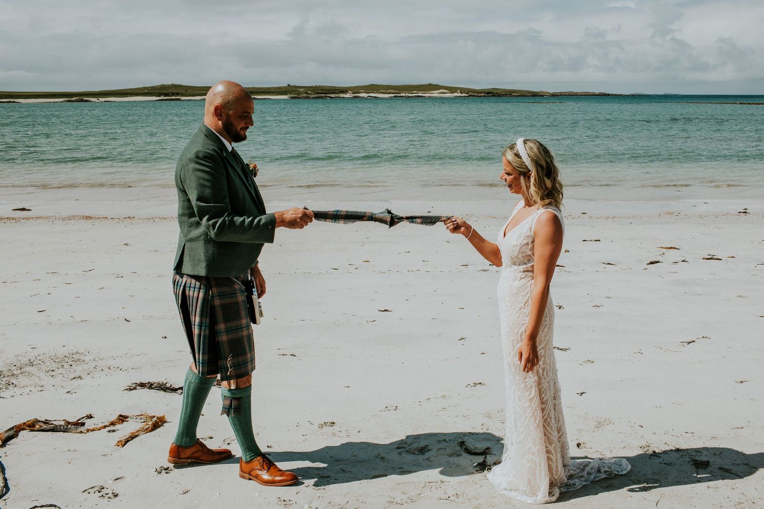 A couple in wedding attire share a moment on a scenic beach with turquoise waters in the background.