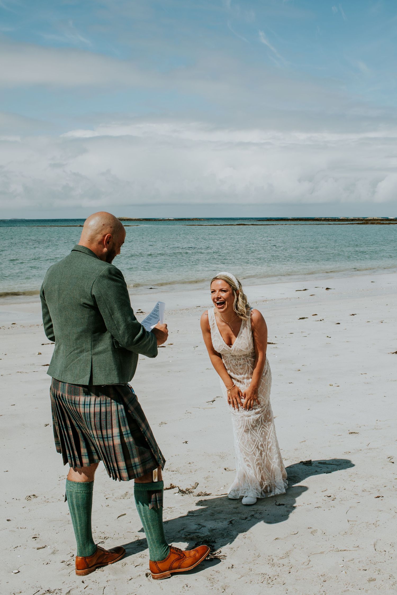 A couple having a beach wedding ceremony in Scotland with the groom wearing a traditional kilt on a sandy shoreline.