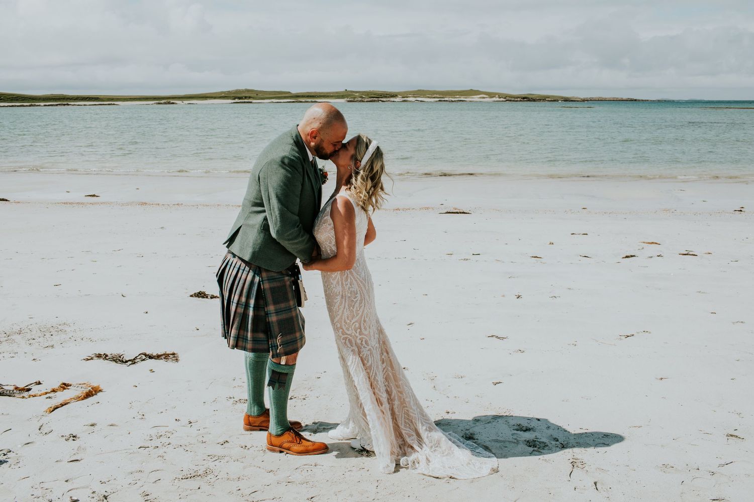 Romantic beach wedding moment with groom in traditional Scottish dress on white sandy shore.