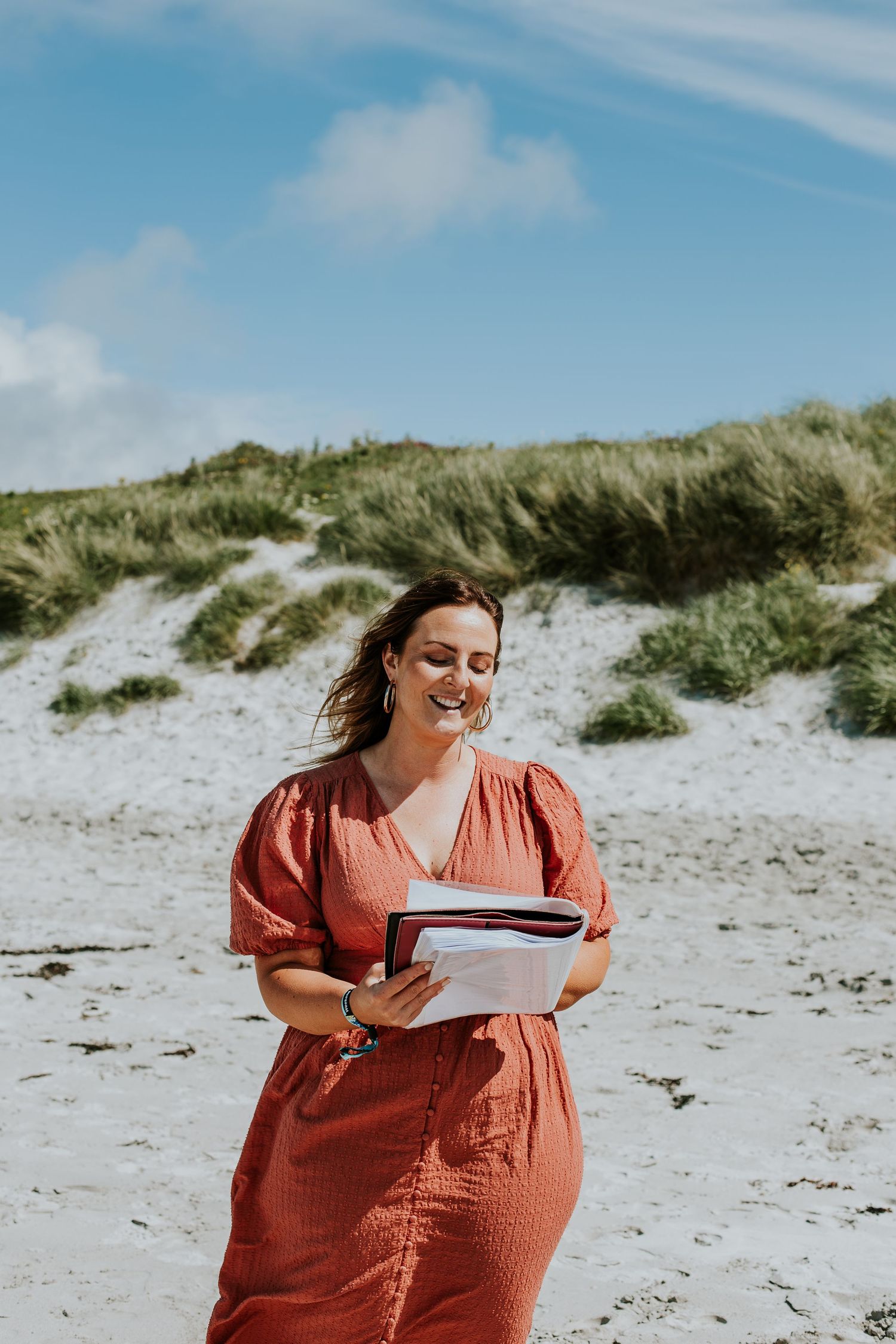 Woman in coral dress smiling while holding papers on scenic beach with dunes.