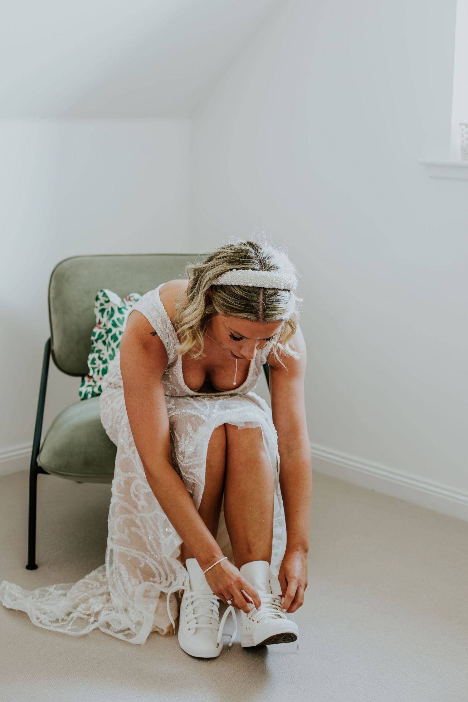 Someone in a white lace dress puts on white sneakers while sitting on a green chair in a bright room.
