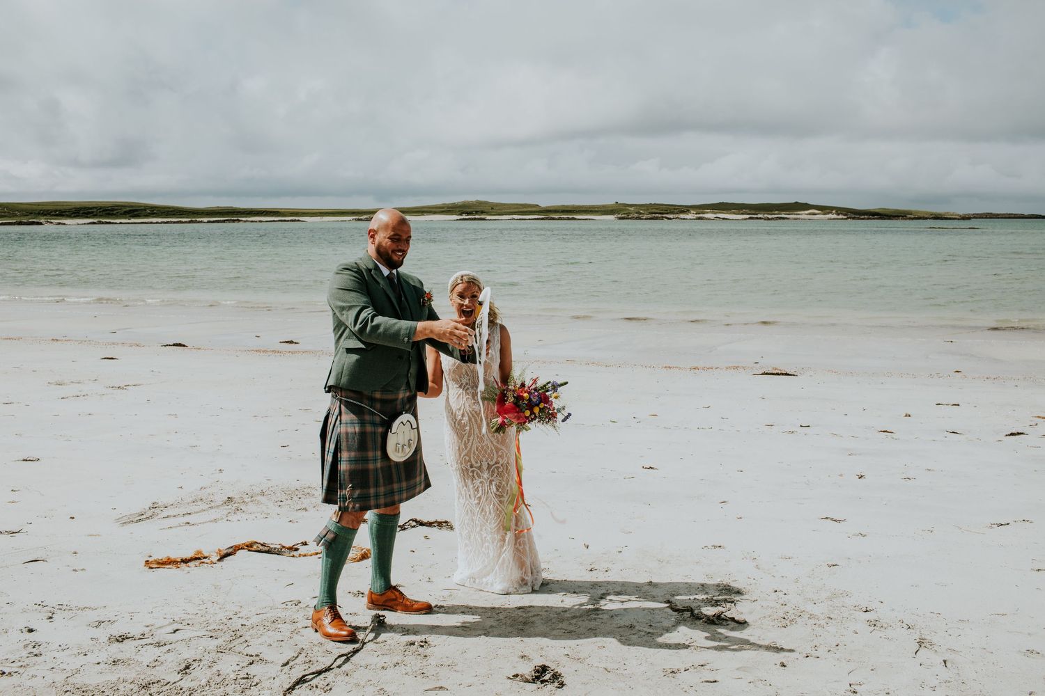 Newlyweds walking together on a pristine white sand beach with traditional Scottish wedding attire.