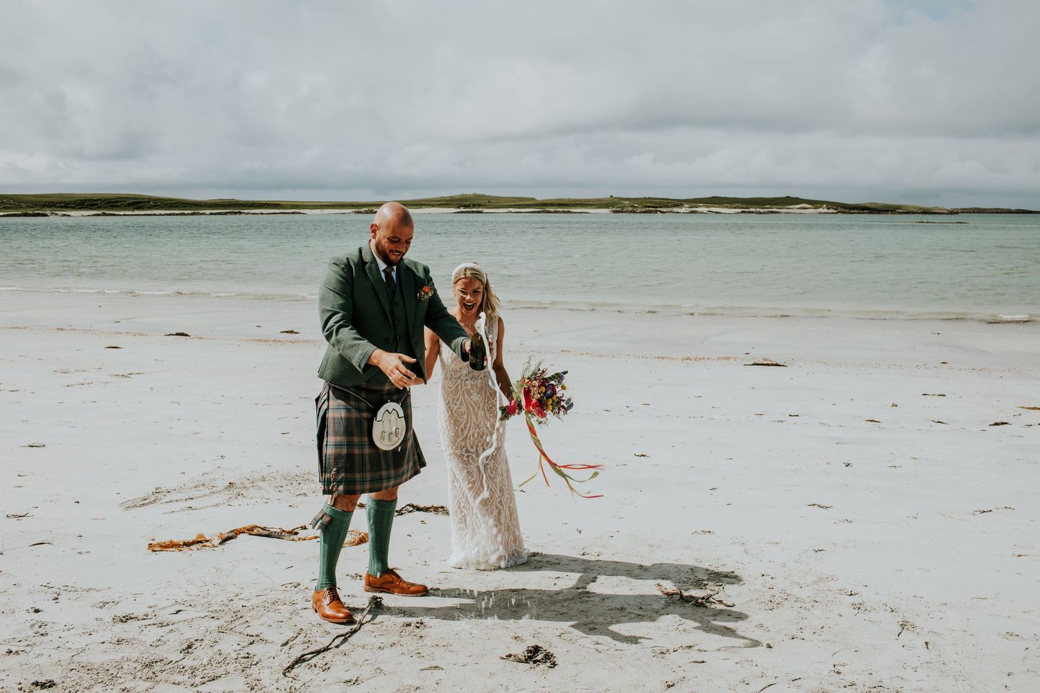 A couple celebrates their wedding day on a serene white sandy beach in Scotland with the groom wearing a traditional kilt.