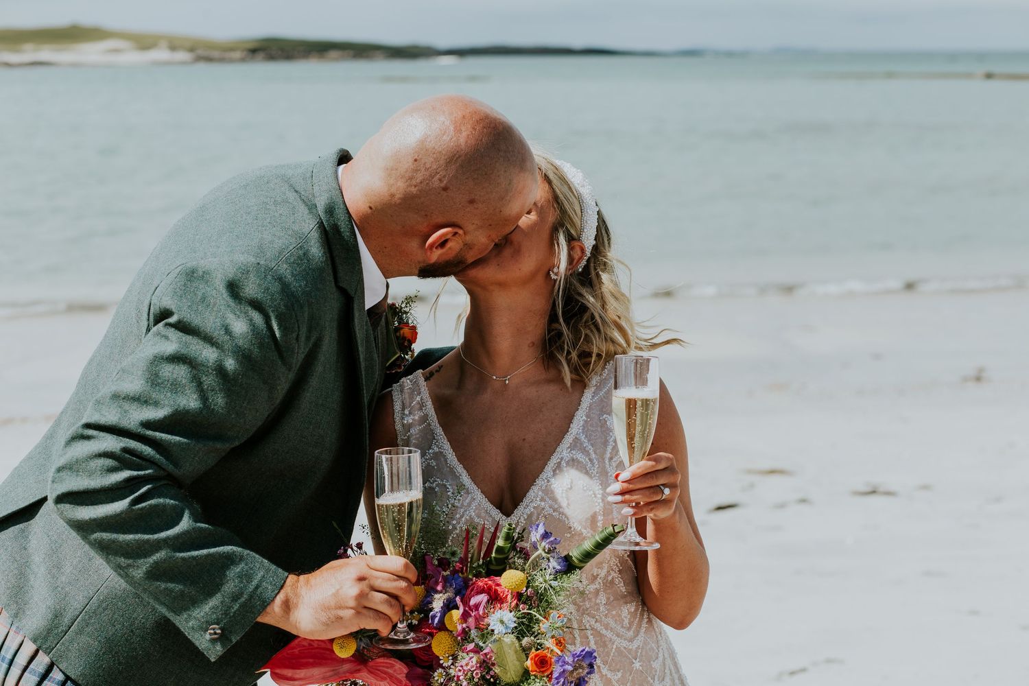 Wedding couple toasting champagne together on beach with colorful bouquet.