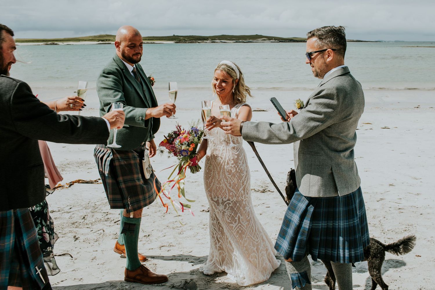 Traditional Scottish wedding celebration takes place on a scenic beach with participants wearing kilts and formal attire.