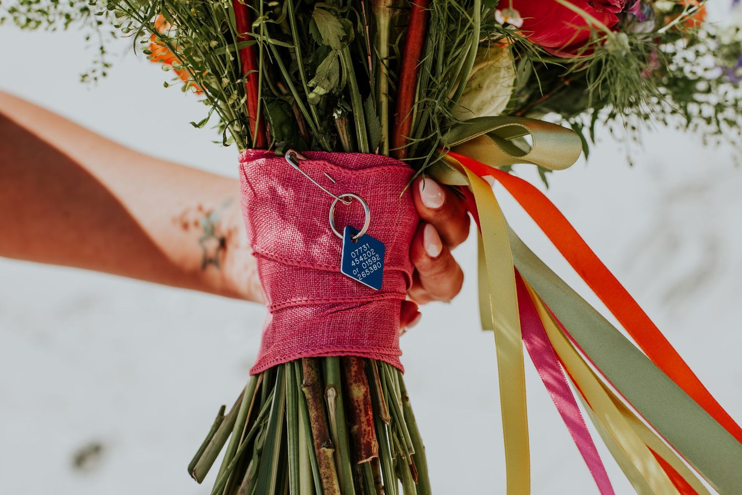 Close-up of hands holding wedding bouquet wrapped with colorful ribbons and blue decorative pin.