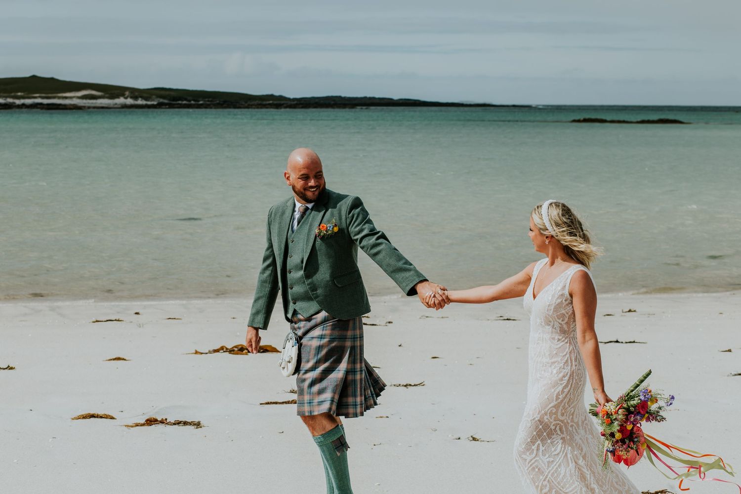 A couple walks hand in hand on a beach during their Scottish-themed wedding celebration.