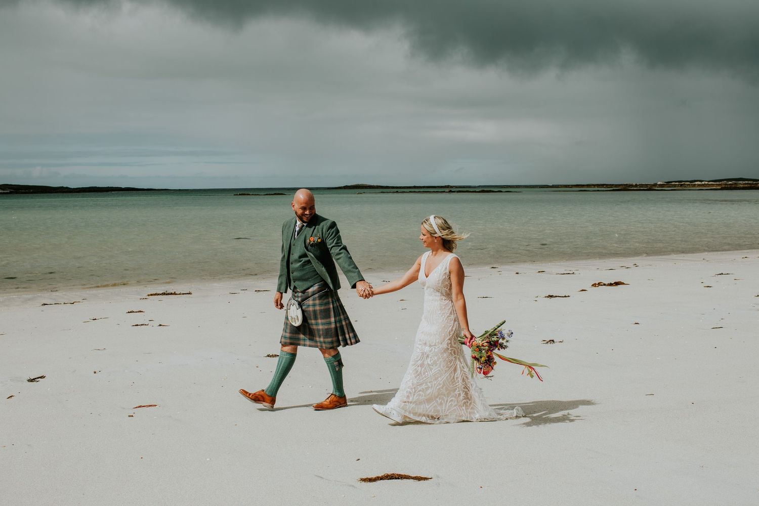 Couple walking hand in hand on white sandy beach under moody sky.