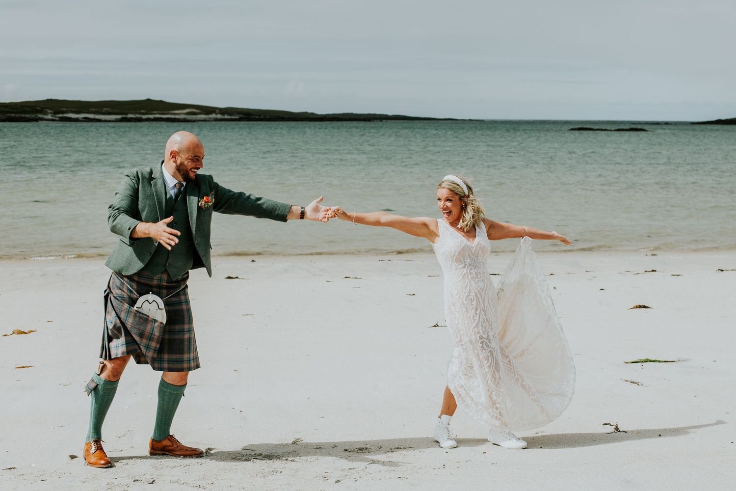 Playful bride and groom in Scottish kilt dancing on beach by ocean.