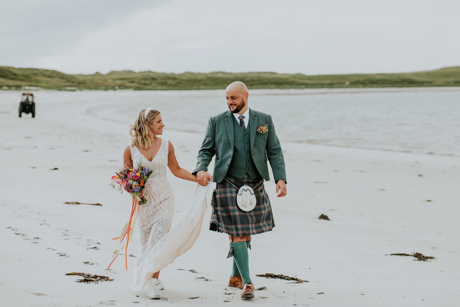 Couple strolling along scenic beach in wedding dress and traditional Scottish kilt.
