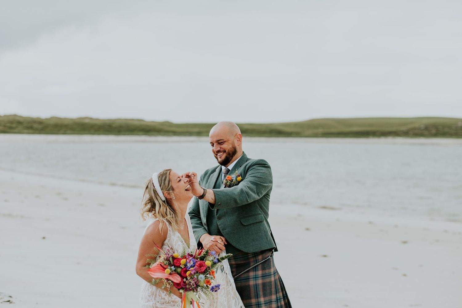 A couple shares a joyful moment by the water on their wedding day, with the bride holding a vibrant bouquet.