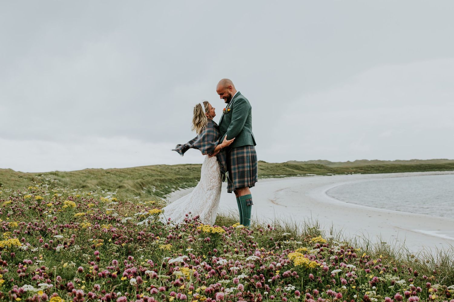 A couple shares an intimate moment on a grassy hillside overlooking a coastal beach in Scotland.