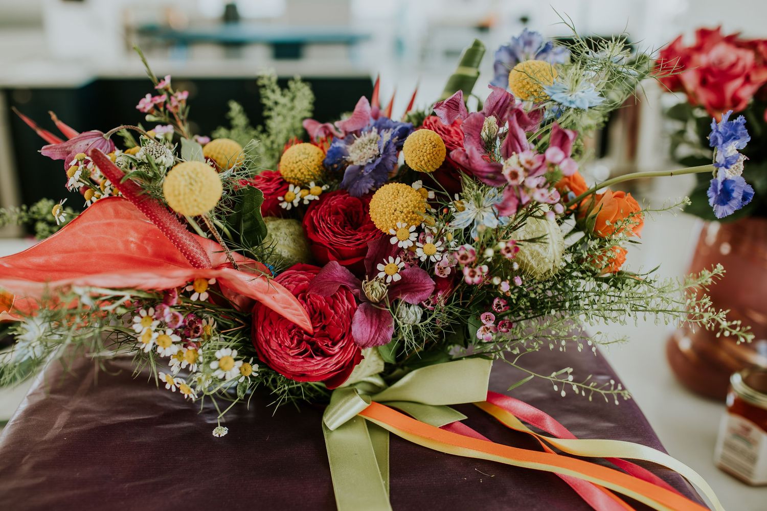 A vibrant floral arrangement with red anthuriums, yellow craspedia, and various colorful wildflowers tied with ribbons.
