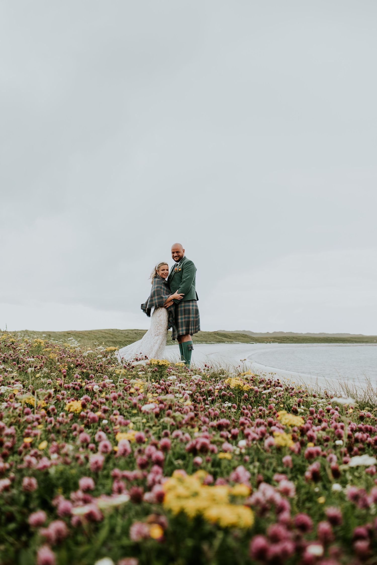 Couple in traditional Scottish attire embraces among colorful wildflowers by a tranquil loch on overcast day.