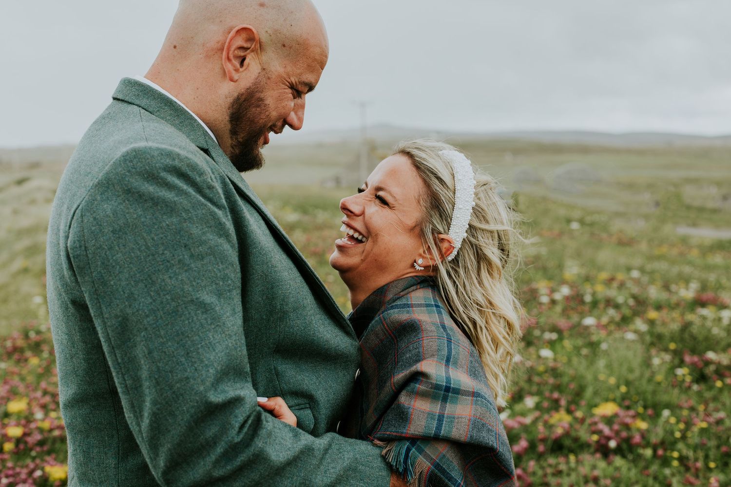 A romantic couple embraces and laughs together on a green hillside wearing coordinating outfits.