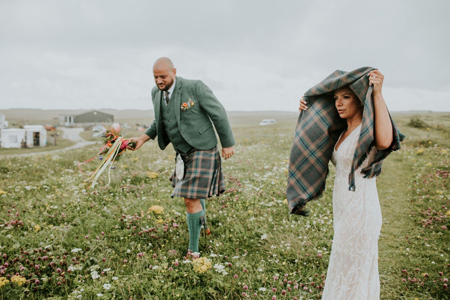 Couple in traditional Scottish attire poses playfully in green meadow with misty mountains in background.