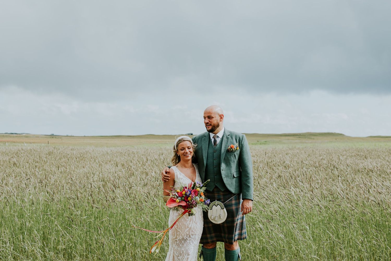 Wedding couple poses in a scenic field wearing traditional Scottish formal wear with a colorful bouquet.