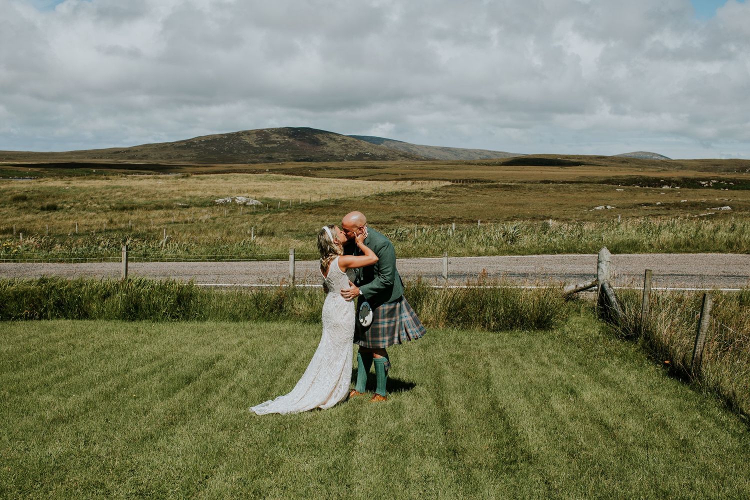 Couple embraces while standing in a grassy meadow with rolling hills and cloudy sky in the Scottish countryside.