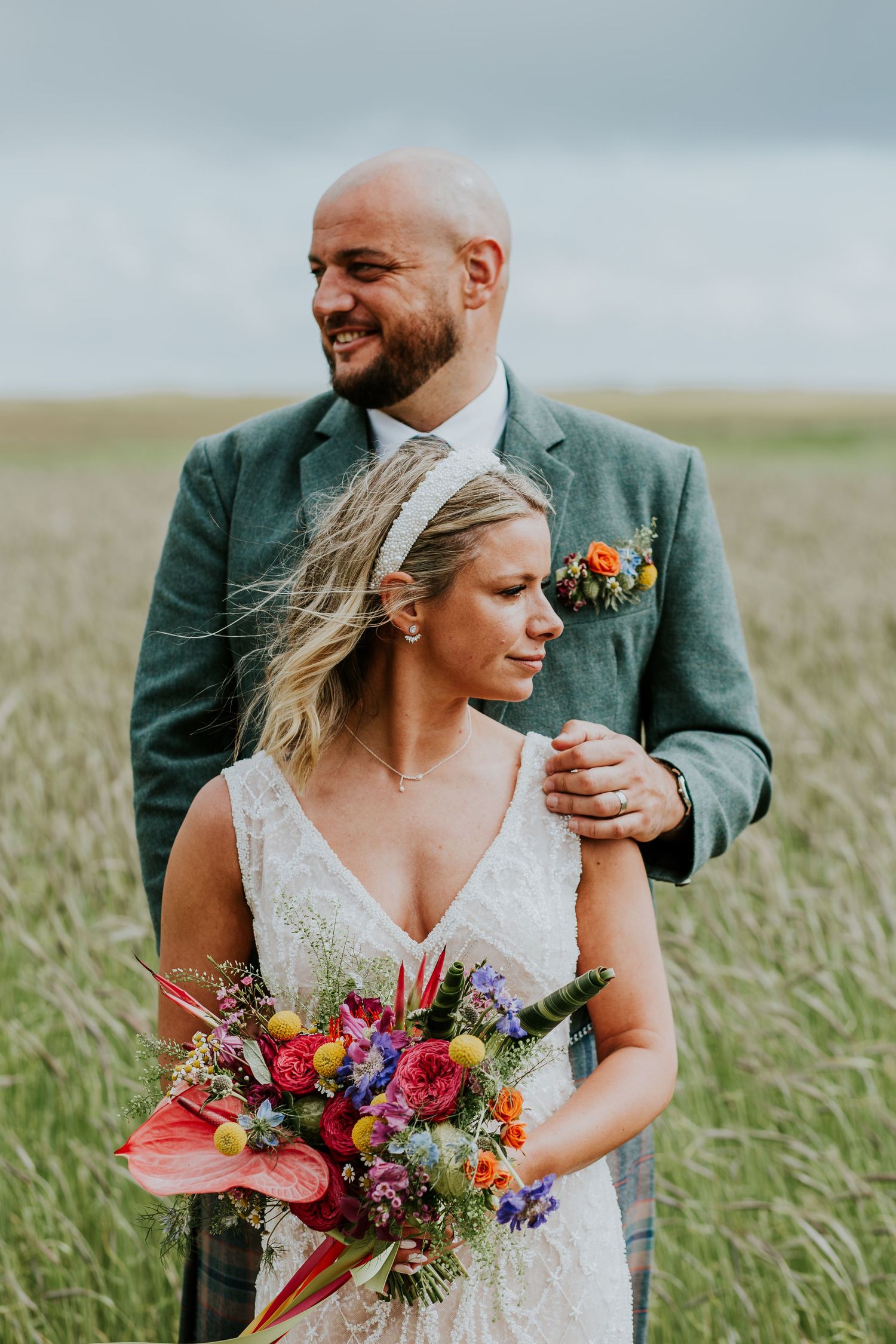 Wedding couple poses intimately in field of tall grass with bride holding vibrant wildflower bouquet.