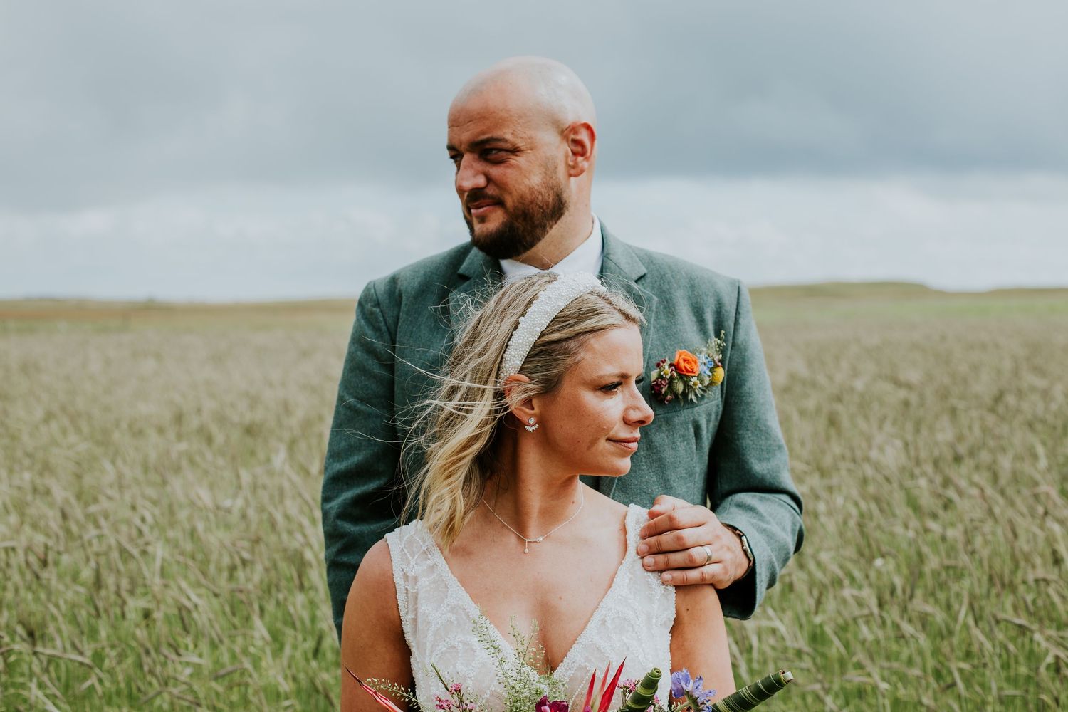 Couple in wedding attire standing in tall grass field under cloudy sky.