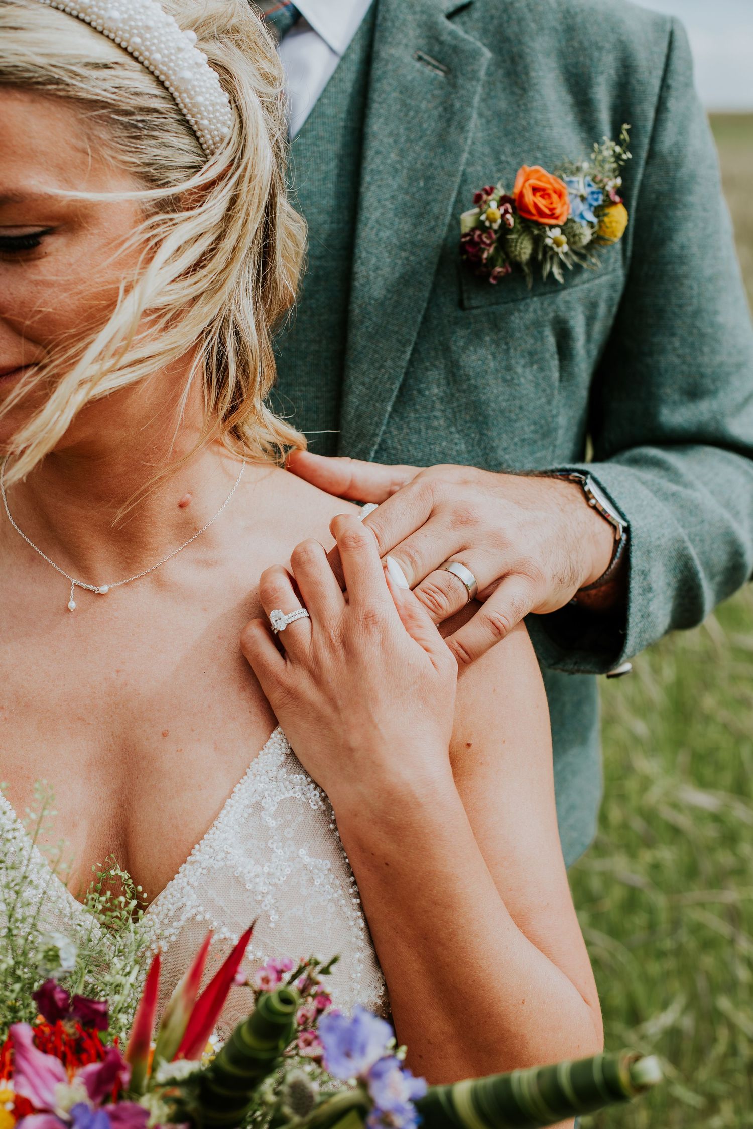 A close-up of hands wearing wedding rings with a green suit jacket and orange boutonniere in view.