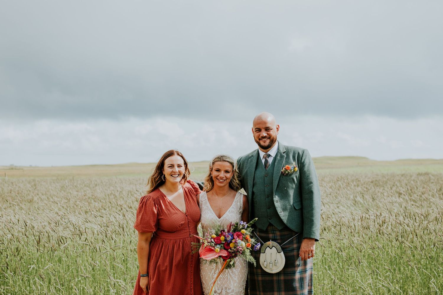 A wedding celebration in a rural Scottish field with traditional Highland dress and bright floral bouquet.
