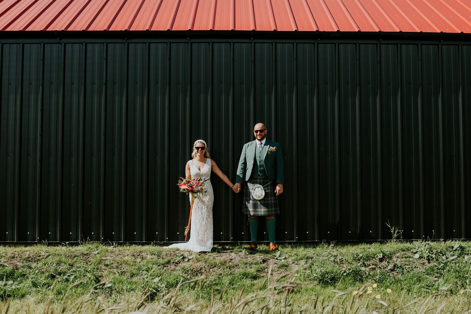 Wedding couple poses against black corrugated metal wall with red roof in Scottish attire.