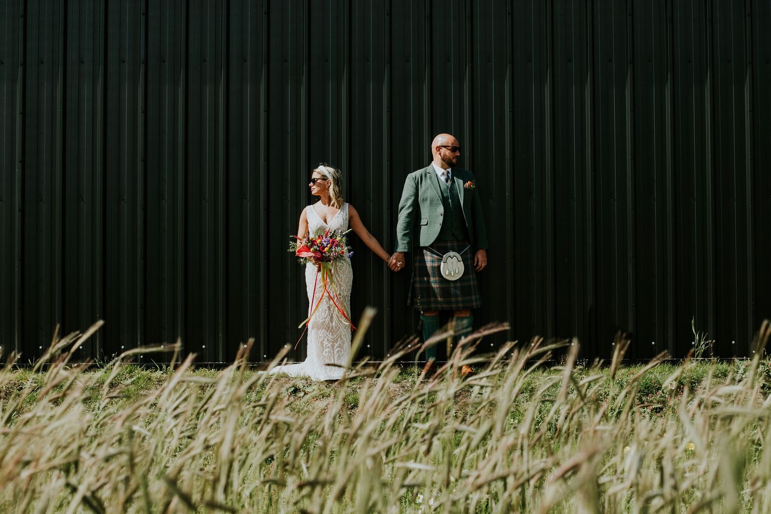 Artistic wedding portrait against black metal wall with tall grass in foreground.
